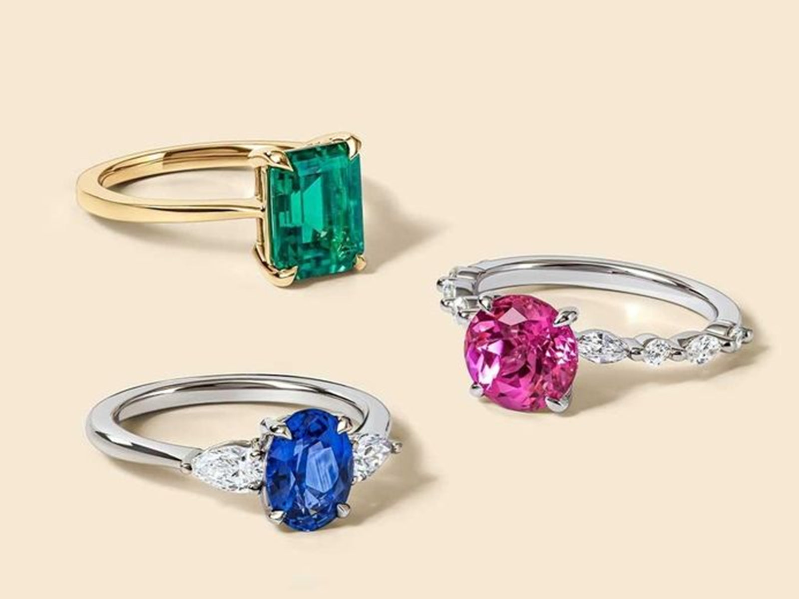 Three colored gemstone rings displayed side by side, including a gold-setting emerald-cut green stone ring, and two silver-setting rings with a blue oval gemstone and a pink round gemstone respectively