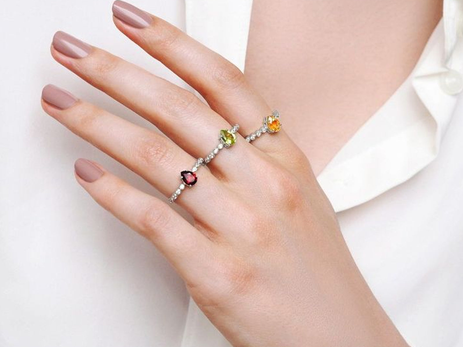 Delicate silver ring with colorful gemstones including peridot, citrine and garnet on slender band worn on manicured hand against white fabric