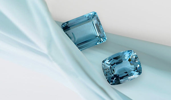 Two stunning loose gemstones, an emerald-cut and a cushion-cut lab-created aquamarine, rest on a soft blue fabric, showcasing the high quality and brilliant color available for lab-created aquamarine rings