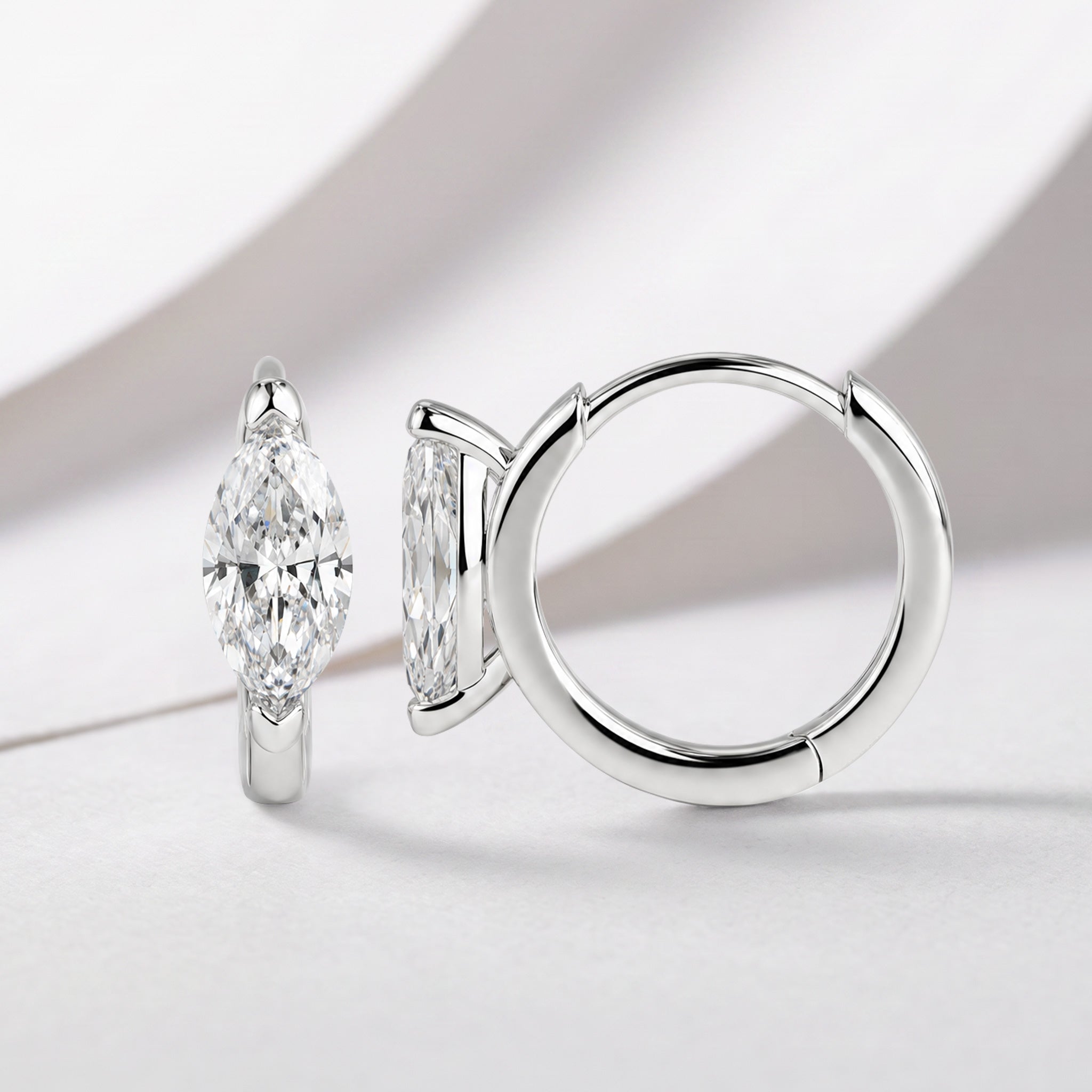 White-gold marquise-shaped hoop earrings displayed on softly draped white fabric.