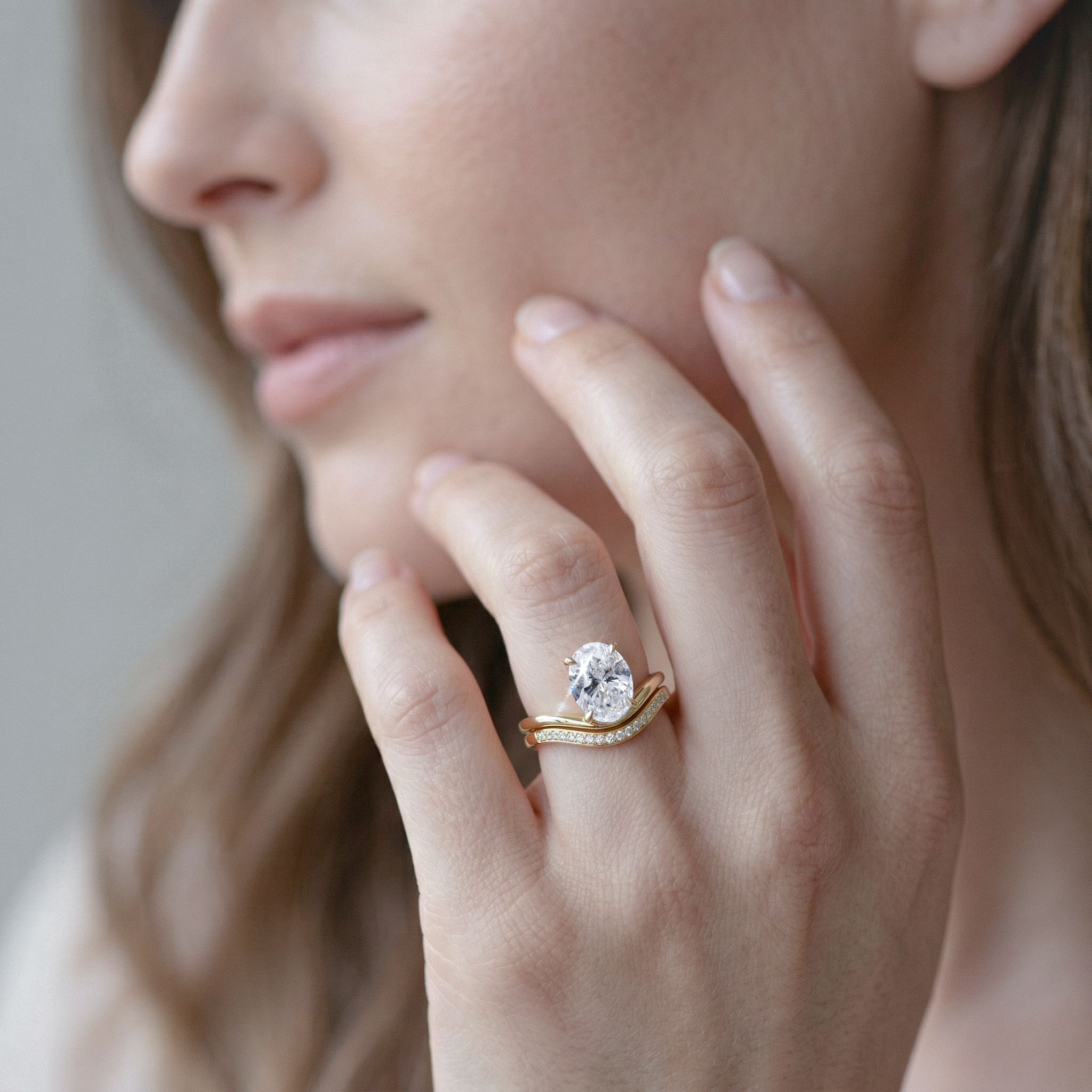 Close-up of a hand wearing gold ring set with oval diamond, touching the face.