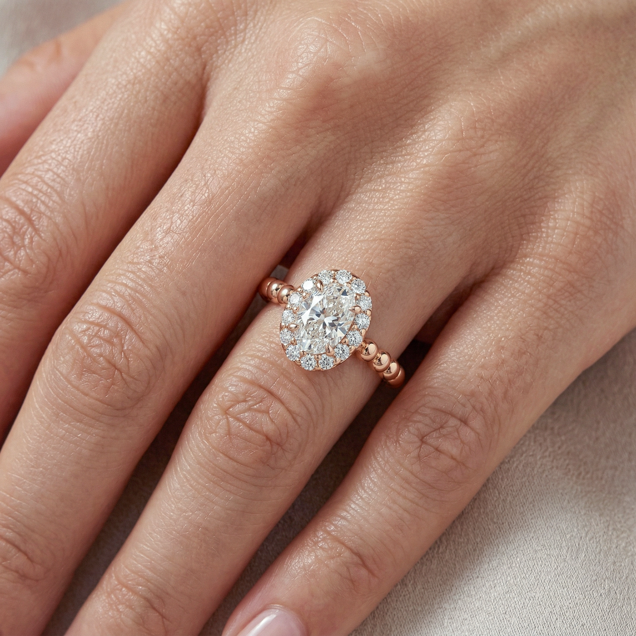 Close-up of an oval halo engagement ring with a rose gold beaded band worn on a hand.