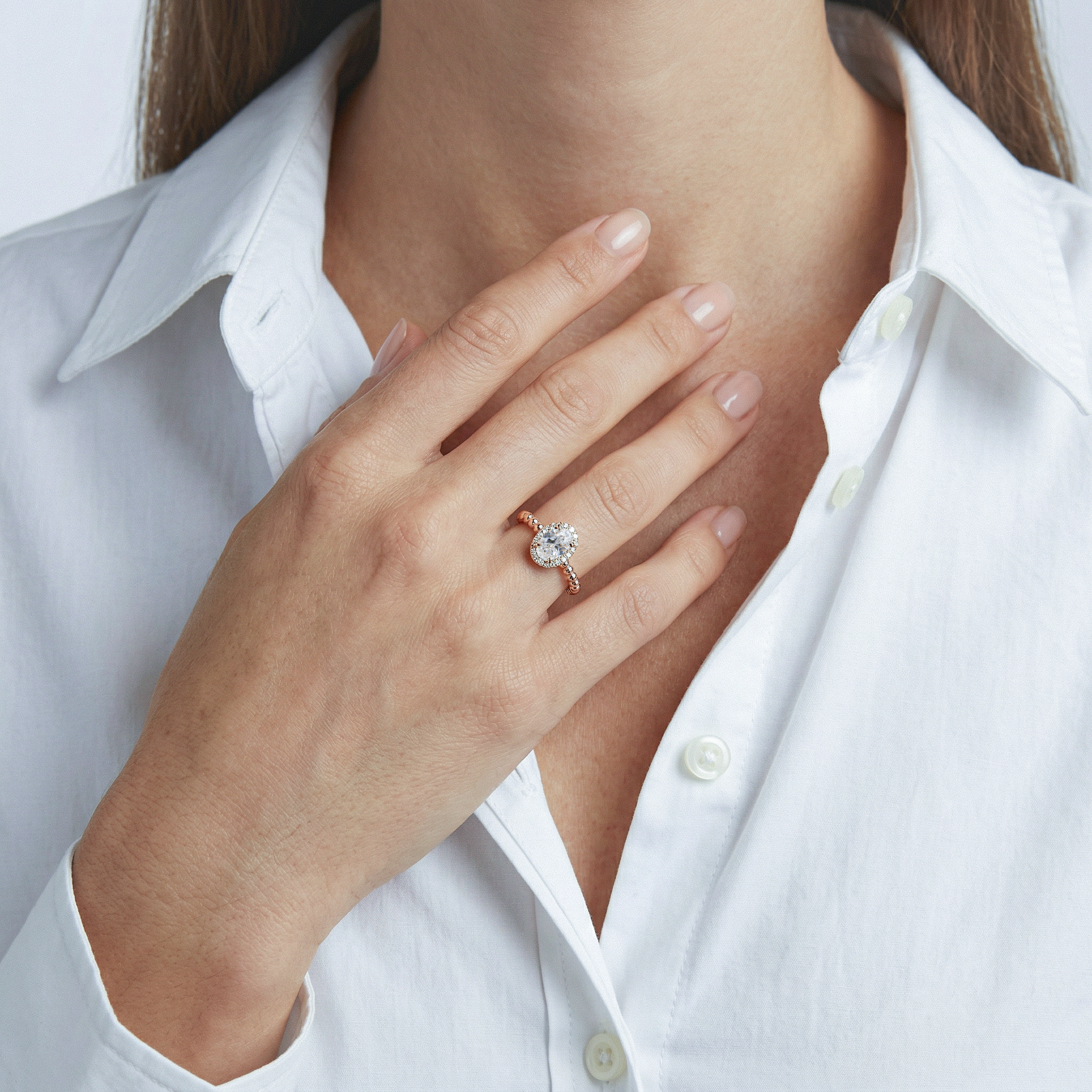 Oval halo engagement ring with a rose gold beaded band worn on a model's hand near her chest.