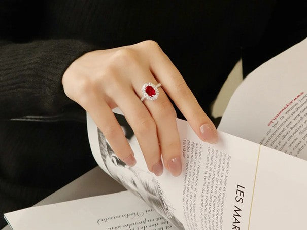 A hand adorned with a stunning lab-grown engagement ring featuring a large red gemstone, while holding a magazine, highlighting the ring's design