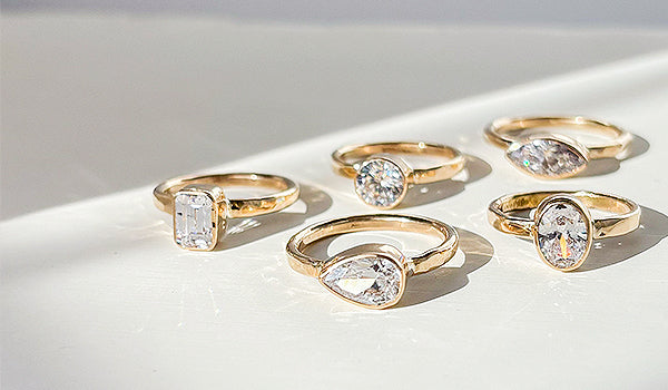 Five gold engagement rings with different diamond cuts and settings arranged on a white surface with soft shadows, showcasing bezel and semi-bezel designs