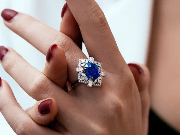 Elegant statement ring featuring a large square-cut blue sapphire gemstone surrounded by a halo of diamonds in a white gold or platinum setting, worn on a woman's hand with burgundy nail polish