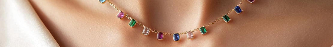 A gold necklace with alternating emerald-cut gemstones in shades of green, pink, blue, and clear, resting on a person's collarbone