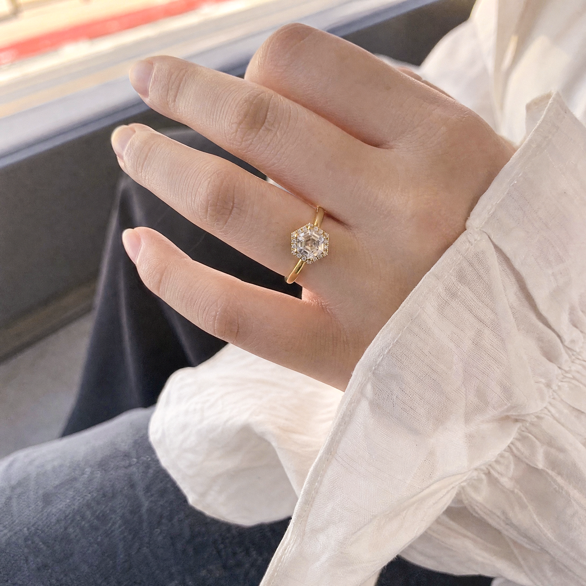Hand wearing a gold ring with a halo hexagon  diamond, sitting on a vehicle seat.