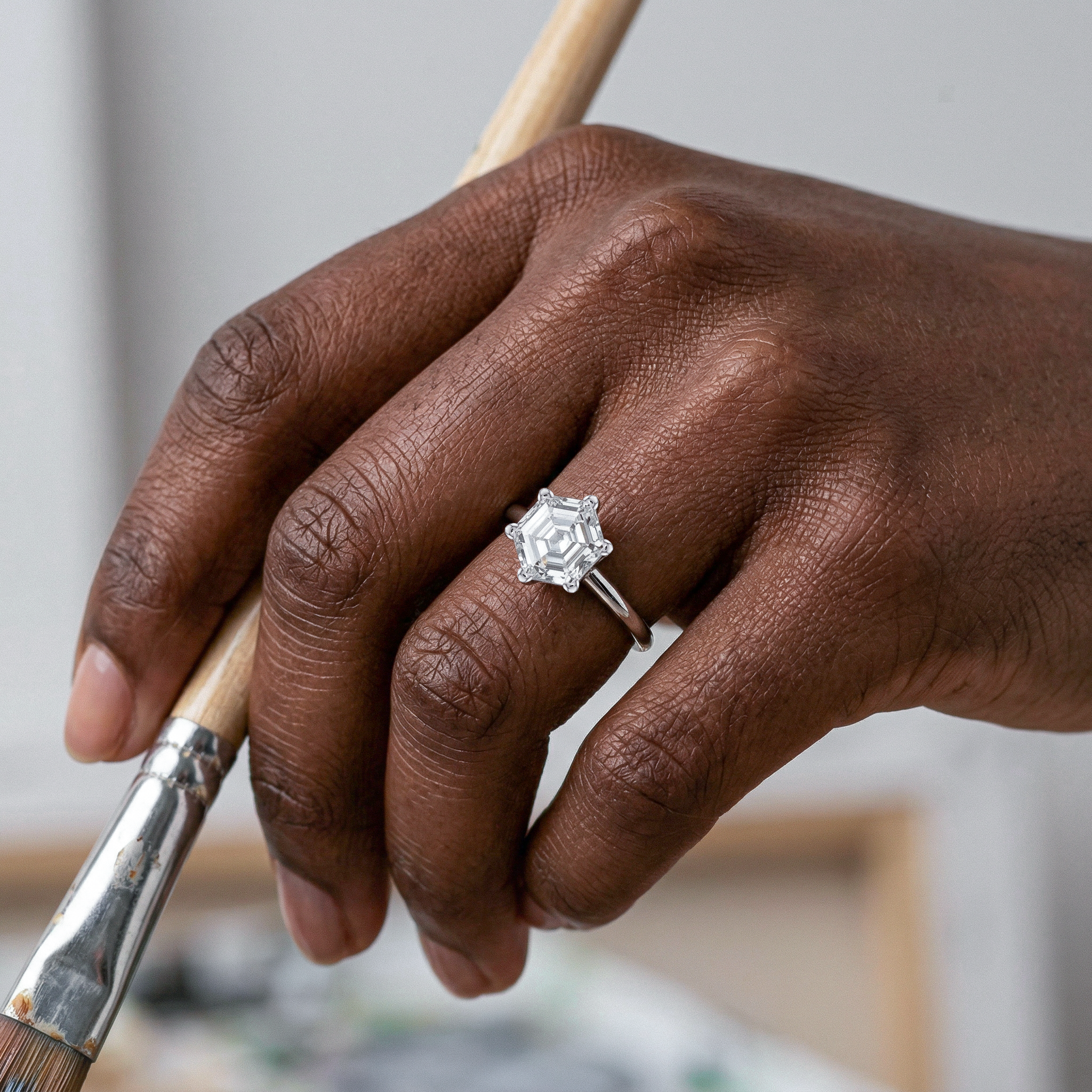 Hand holding a paintbrush with a hexagon diamond ring on a blurred background