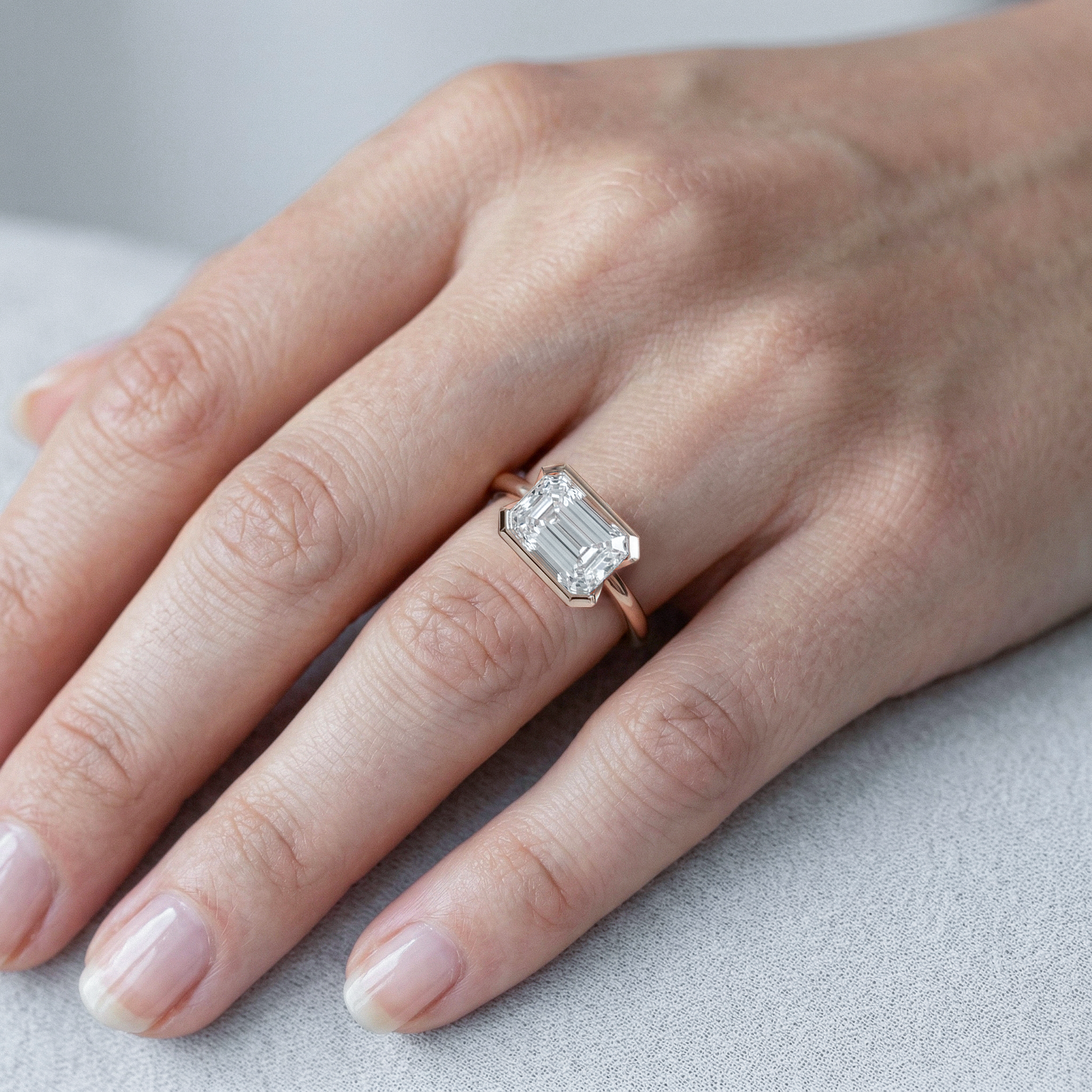 Hand wearing a rose gold ring with a emerald cut diamond on a light gray background