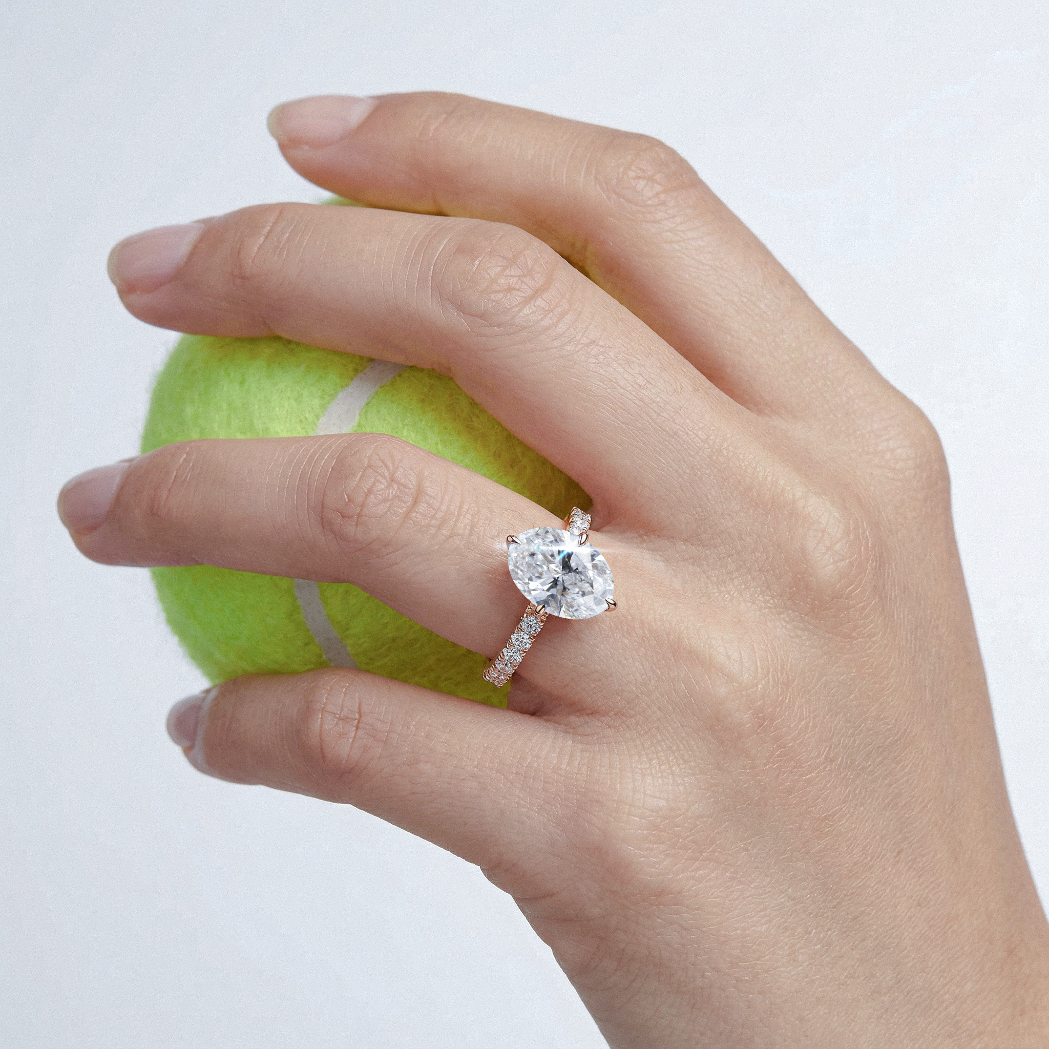 Hand wearing a oval diamond pave ring holding a tennis ball on a white background