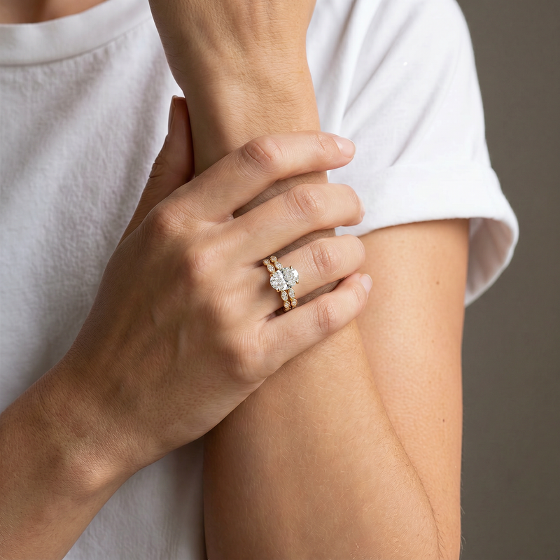 A close-up of a person's arm and hand wearing a gold ring with a clear oval -cut center stone and small side stones, paired with a white t-shirt against a neutral gray background.