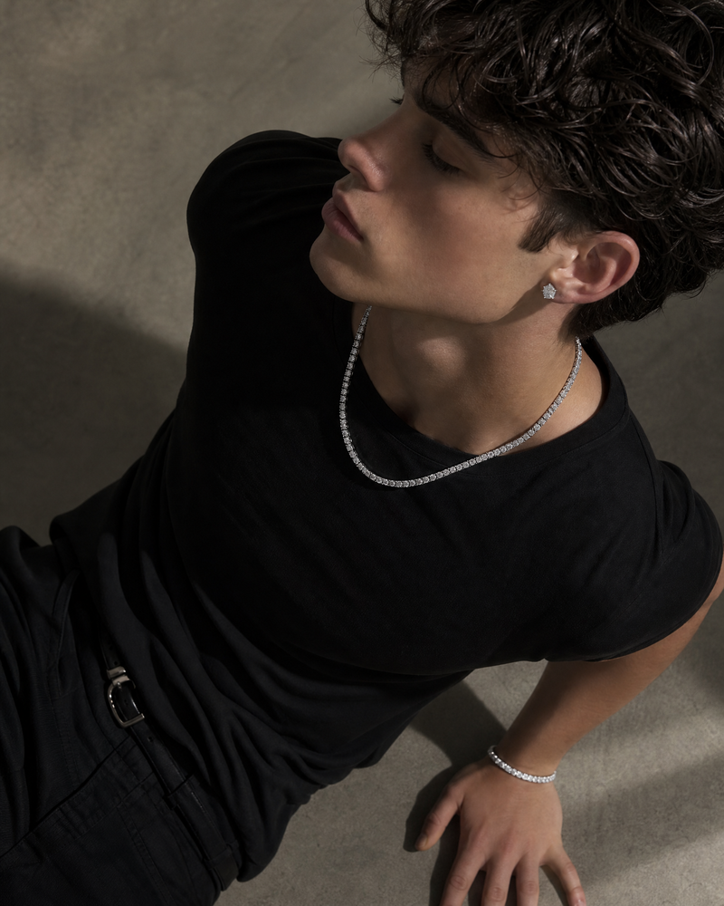 A man wearing a white gold  diamond-encrusted chain necklace and a matching  bracelet, styled with a black t-shirt and dark jeans against a textured concrete background.